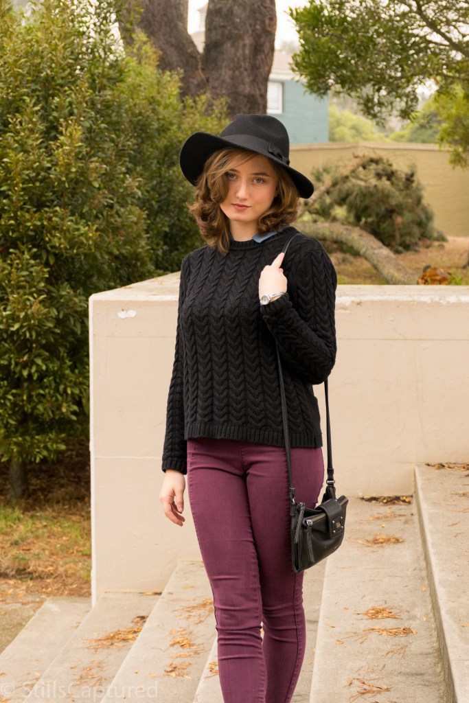 Black hat, black sweater with blue collar, eggplant pants, black booties, long bob, cross body purse